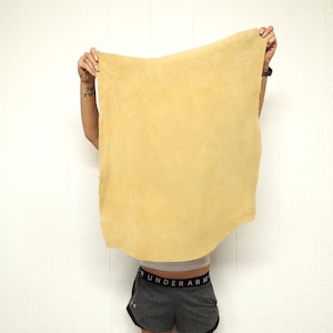 May include: A large, rectangular piece of tan-colored leather is held up against a white wall. The leather has a soft, textured appearance. The image focuses on the leather, showcasing its natural color and surface.