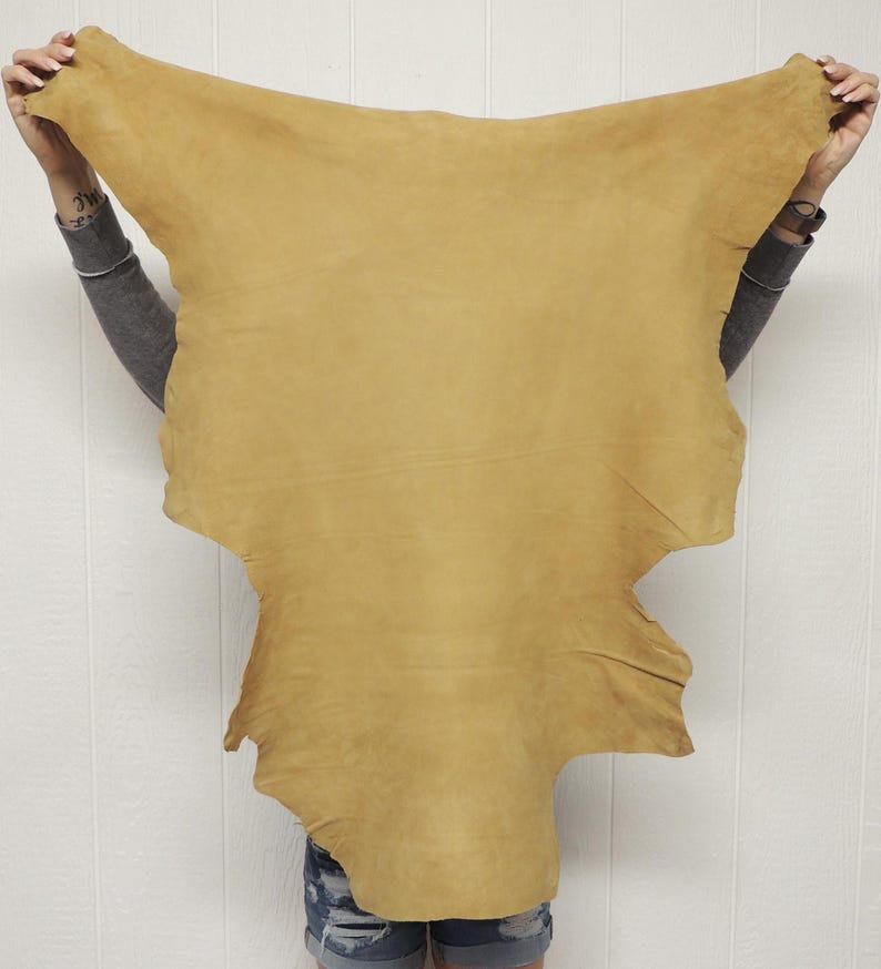 May include: A large, irregularly shaped piece of tan-colored leather hide with a smooth, matte finish. The material features natural variations in tone and texture across its surface, displaying a soft, flexible appearance throughout.