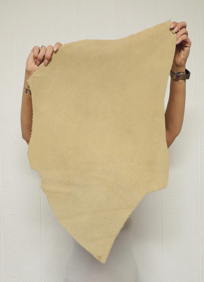 May include: A piece of tan-colored leather with a textured surface. The leather is held up by a person, showcasing its irregular shape and soft appearance. The background is a white wall.