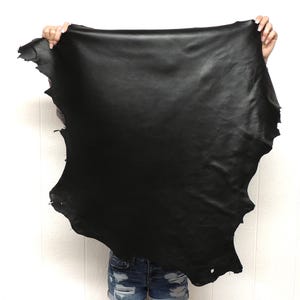 May include: A large, irregularly shaped piece of black leather is held up against a white background. The leather has a smooth, matte finish and appears to be supple. The edges are raw and uneven, suggesting it is a hide.