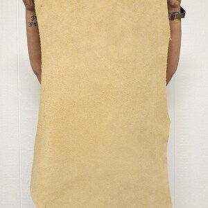 May include: A large, rectangular piece of tan-colored leather is held up against a white background. The leather has a rough, textured surface and appears to be unfinished. The edges are uneven, suggesting it's been cut from a larger hide. The leather is held up by a person.