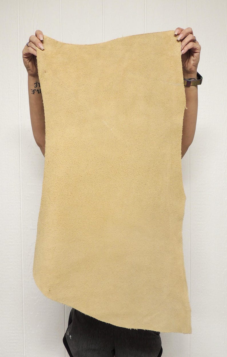May include: A large, rectangular piece of tan-colored leather is held up against a white background. The leather has a rough, textured surface and appears to be unfinished. The edges are uneven, suggesting it's been cut from a larger hide. The leather is held up by a person.