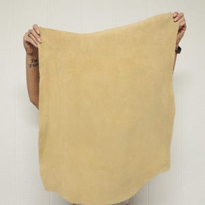 May include: A large, rectangular piece of tan-colored leather is held up against a white background. The leather has a rough, textured surface and appears to be flexible. The edges are uneven, suggesting it's been cut from a larger hide. The leather is likely for crafting.