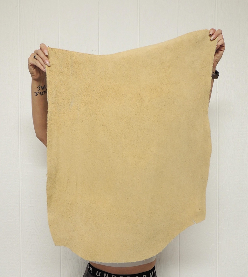 May include: A large, rectangular piece of tan-colored leather is held up against a white background. The leather has a rough, textured surface and appears to be flexible. The edges are uneven, suggesting it's been cut from a larger hide. The leather is likely for crafting.