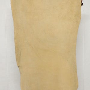 May include: A large, rectangular piece of tan-colored leather is held up against a white background. The leather has a rough, textured surface and appears to be a single, uncut hide. The edges are uneven, suggesting a natural, raw material.
