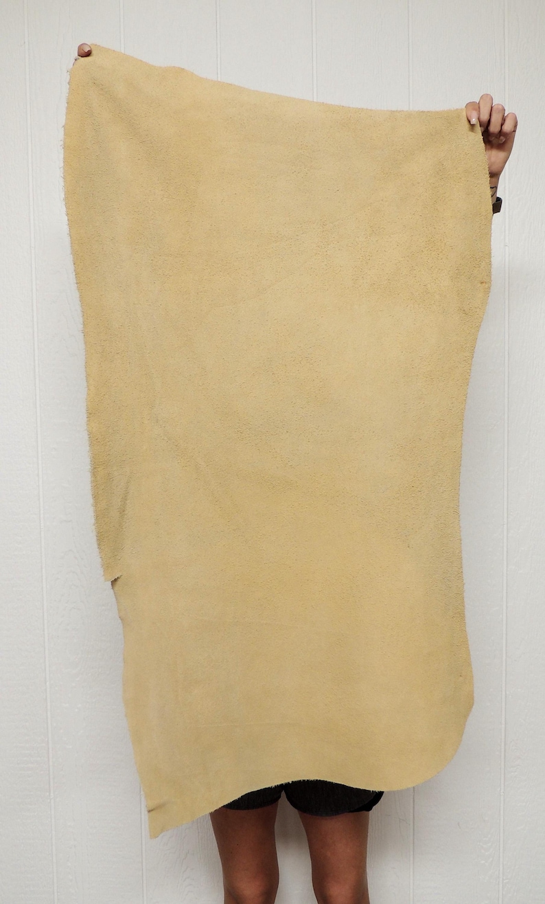 May include: A large, rectangular piece of tan-colored leather is held up against a white background. The leather has a rough, textured surface and appears to be a single, uncut hide. The edges are uneven, suggesting a natural, raw material.