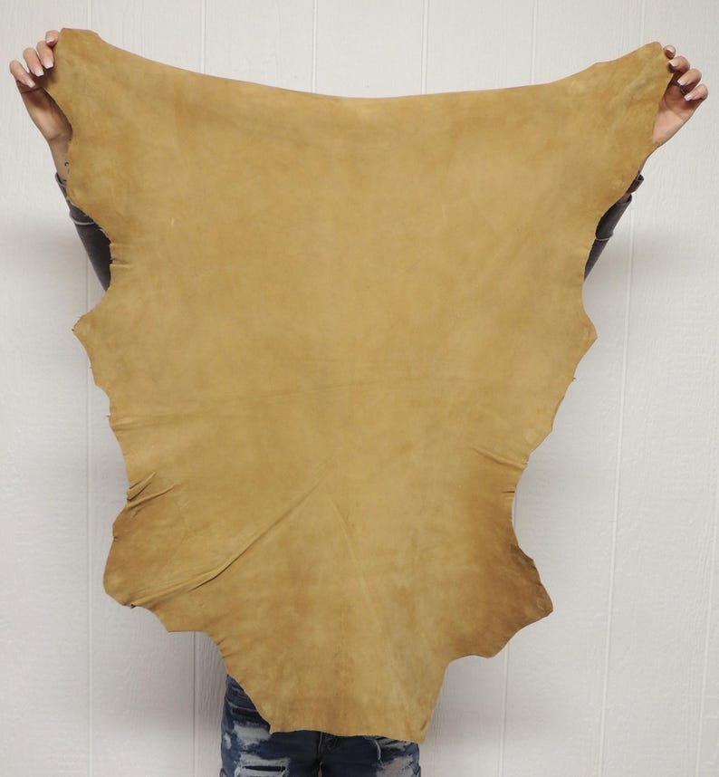 May include: A large, irregularly shaped piece of tan-colored suede leather with a soft, matte texture. The hide features natural variations in tone and a smooth, flexible surface, displaying the organic edges characteristic of raw animal skin.