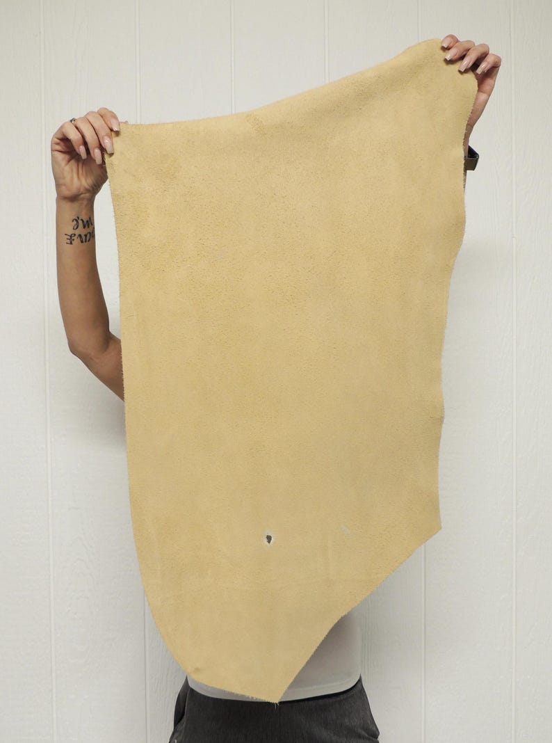 May include: A piece of tan-colored leather is held up against a white background. The leather has a slightly irregular shape and a visible texture. A small hole is present near the bottom. The leather is being held up by a person.