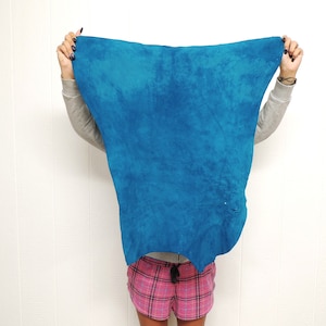 May include: A person is holding a large, blue piece of fabric in front of their face. The fabric has a mottled, uneven texture. The person is wearing a pink and black plaid skirt.