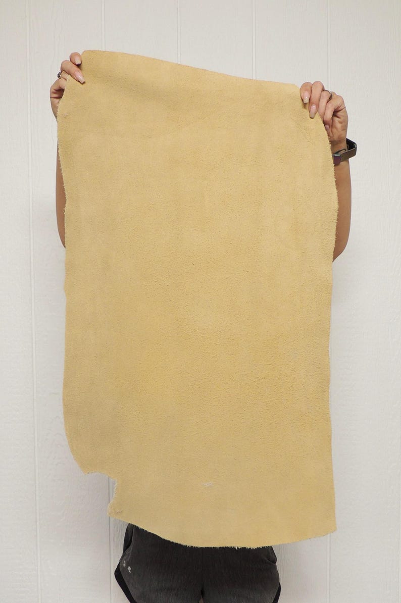 May include: A large, irregularly shaped piece of tan-colored leather. The leather has a soft, suede-like texture and is held up against a white wall. The edges of the leather are uneven, suggesting it is a natural cut.