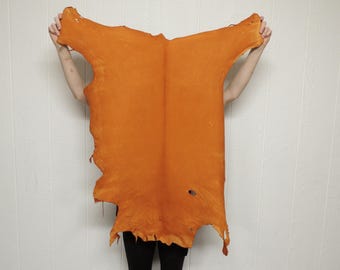 May include: A large, irregular piece of orange-colored leather hide with a smooth, matte finish. The hide features natural edges, a visible central spine line, and a small circular hole near the bottom right section of the material surface.