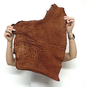 May include: A large, irregularly shaped piece of medium brown leather with a distinct, deeply wrinkled and pebbled texture. The hide features natural edges and a consistent, rich tan color throughout its surface, showcasing a rugged grain pattern.