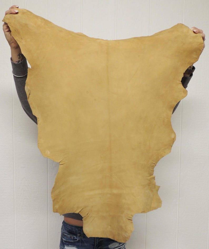May include: A large, irregularly shaped piece of tan-colored leather hide with a smooth, matte surface. The hide features natural edges and a consistent light brown tone across its entire surface, displaying the organic texture of the material.