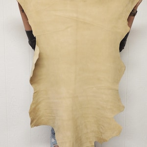 May include: A large, irregularly shaped piece of tanned leather, held up against a white background. The leather is a light tan color with a slightly textured surface. The edges are uneven, suggesting it's been cut from a larger hide. The overall shape is organic.