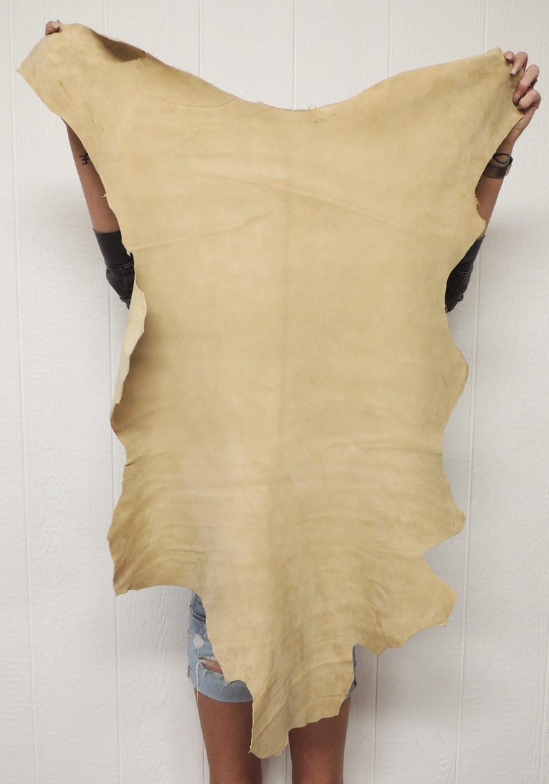 May include: A large, irregularly shaped piece of tanned leather, held up against a white background. The leather is a light tan color with a slightly textured surface. The edges are uneven, suggesting it's been cut from a larger hide. The overall shape is organic.