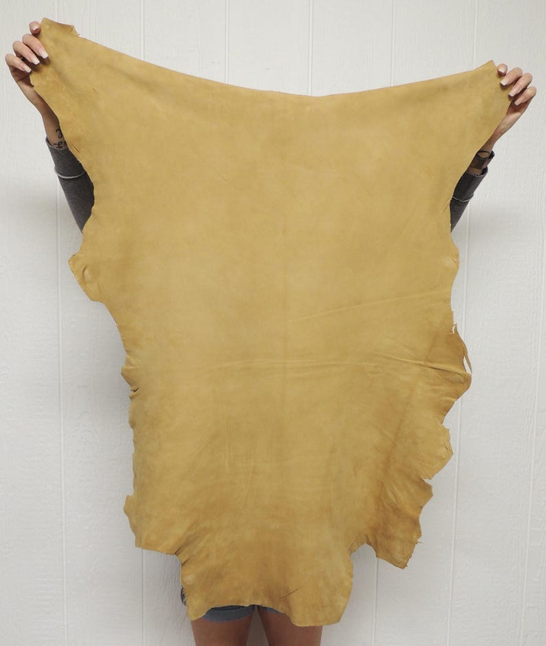 May include: A large, irregularly shaped piece of tan-colored leather hide with a smooth, matte finish. The material features natural edges and a consistent, warm golden-brown tone across its entire surface, displaying a soft, pliable texture.
