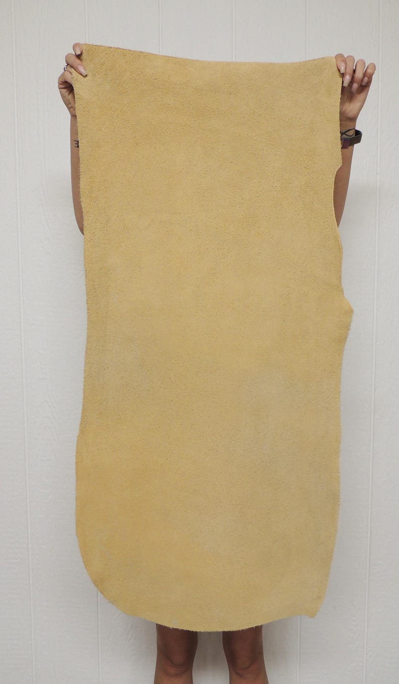 May include: A large, rectangular piece of tan-colored leather with a textured surface. The leather is held up by a person, showcasing its size and shape. The edges are slightly irregular, indicating a natural cut.
