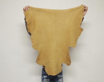 May include: A large, irregularly shaped piece of tan-colored leather hide with a smooth, matte surface. The hide features natural, organic edges and a consistent light brown tone throughout, displaying the raw texture of the material.
