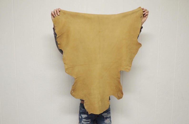 May include: A large, irregularly shaped piece of tan-colored leather hide with a smooth, matte surface. The hide features natural, organic edges and a consistent light brown tone throughout, displaying the raw texture of the material.