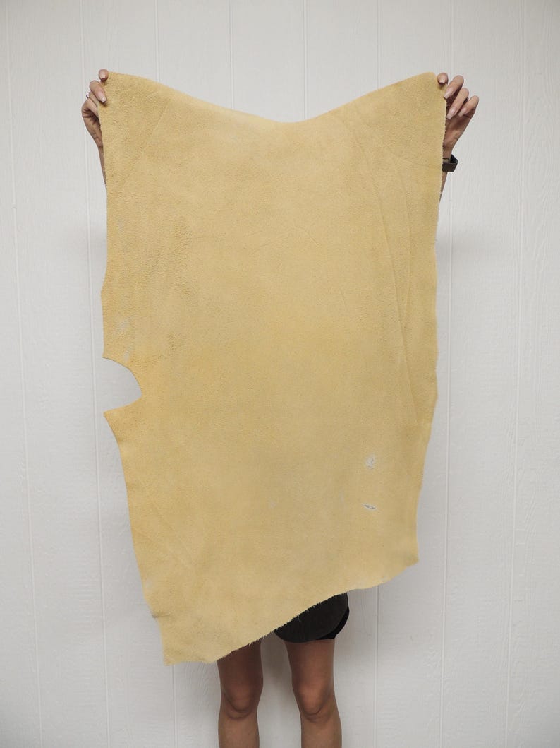 May include: A large, irregularly shaped piece of tan-colored leather is held up by two hands. The leather has a soft, suede-like texture and a few small imperfections. The edges are uneven, and there is a cutout on the left side.