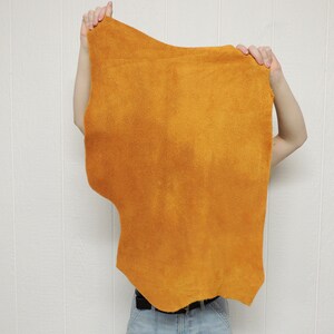 May include: A large piece of orange leather, held up by a person's hands. The leather is soft and supple, with a slightly textured surface.