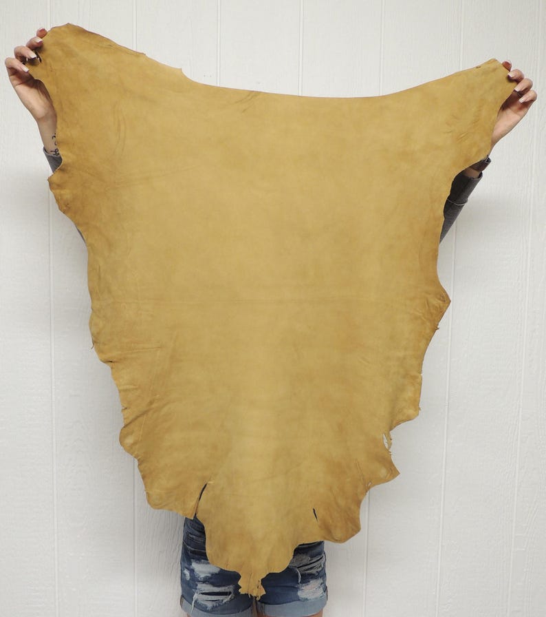May include: A large, irregularly shaped piece of tan-colored leather hide with a smooth, matte finish. The hide features natural edges and a consistent, warm golden-brown tone across its entire surface, displaying a soft, pliable texture.