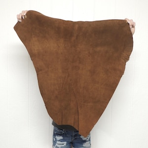 May include: A large, irregularly shaped piece of brown leather is held up against a white background. The leather has a textured surface and a rich, warm brown color. The edges of the leather are slightly uneven.