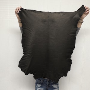 May include: A large, irregularly shaped piece of black leather is held up against a white background. The leather has a textured surface and appears to be a single hide. The edges are uneven, suggesting it is raw and unprocessed. The image focuses on the leather's color and texture.