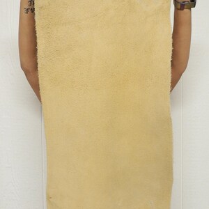 May include: A large, rectangular piece of tan-colored leather is held up against a white background. The leather has a soft, suede-like texture and appears to be a single, uncut hide. The edges are slightly uneven, suggesting a natural cut.