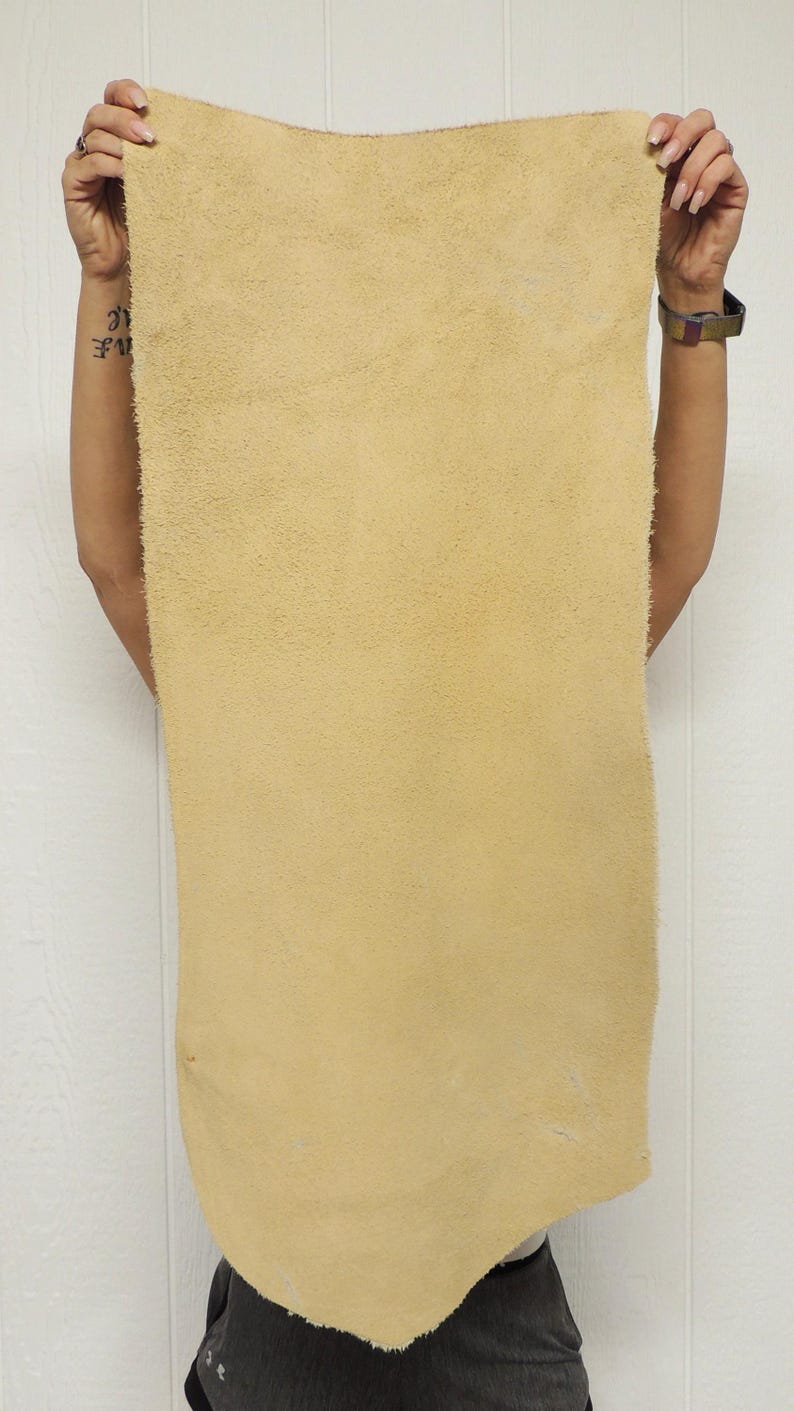 May include: A large, rectangular piece of tan-colored leather is held up against a white background. The leather has a soft, suede-like texture and appears to be a single, uncut hide. The edges are slightly uneven, suggesting a natural cut.