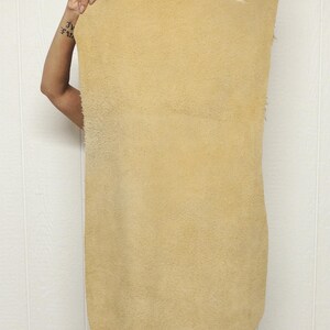 May include: A large, rectangular piece of tan-colored leather is held up against a white wall. The leather has a rough, textured surface and a natural, organic appearance. The edges are uneven.