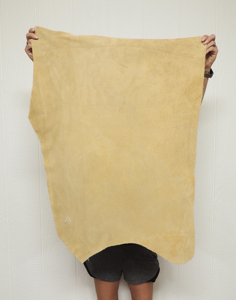 May include: A large, irregularly shaped piece of tan-colored leather is held up against a white background. The leather has a soft, suede-like texture and appears to be a single hide. The edges are uneven, showing the natural shape of the material.