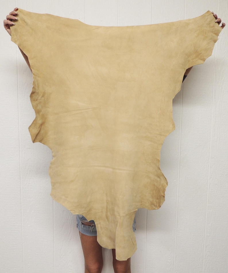 May include: A large, irregularly shaped piece of tan leather is held up against a white background. The leather has a soft, suede-like texture and varying shades of tan. The edges are uneven, suggesting it's a natural hide.