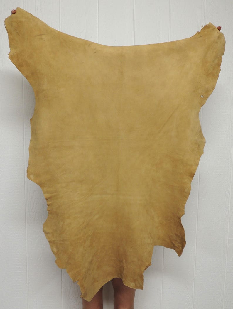 May include: A large, irregularly shaped piece of tan-colored leather hide with a smooth, matte surface. The material features natural variations in tone and texture across its surface, with raw, uneven edges along the entire perimeter.