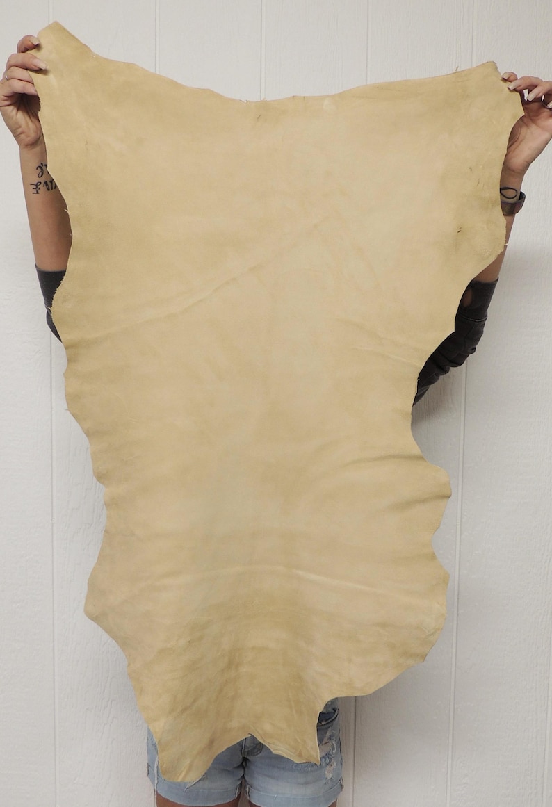May include: A large, irregularly shaped piece of tan-colored leather. The leather is held up against a white background, showcasing its natural texture and color variations. The hide appears to be soft and pliable.