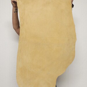 May include: A large, irregularly shaped piece of tan leather is held up against a white background. The leather has a soft, suede-like texture and a natural, organic shape. The color is a warm, muted yellow-brown.