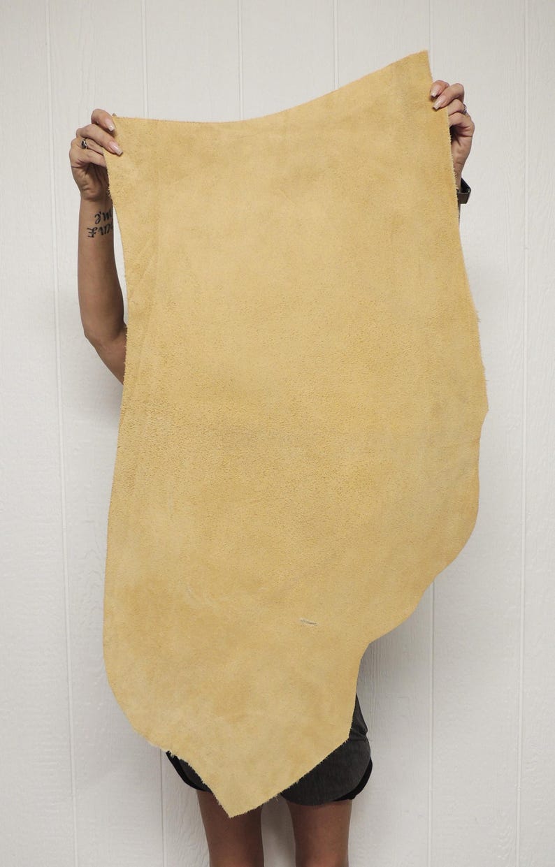 May include: A large, irregularly shaped piece of tan leather is held up against a white background. The leather has a soft, suede-like texture and a natural, organic shape. The color is a warm, muted yellow-brown.