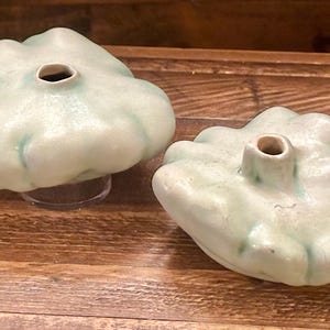 May include: Two light green ceramic objects shaped like a pattypan squash, each with a central hole. The objects have a slightly textured surface and sit on a wooden surface. The objects are likely decorative or functional.