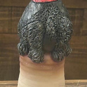 May include: A ceramic dog figurine with a black body and a red collar. The dog has a textured, shaggy appearance and is perched atop a beige and brown cylindrical base. The figurine is a decorative item.