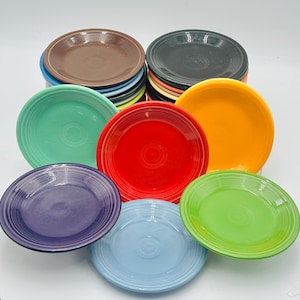 May include: A collection of colorful ceramic plates in various shades, including red, orange, green, blue, purple, brown, and black. The plates are stacked and arranged, showcasing their vibrant hues and classic design. These plates are ideal for serving food.