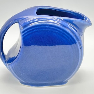 May include: A cobalt blue ceramic pitcher with a large handle and a spout. The pitcher has a rounded body and a smooth, glossy finish. The handle is a large, open loop. The pitcher is approximately 8 inches tall.