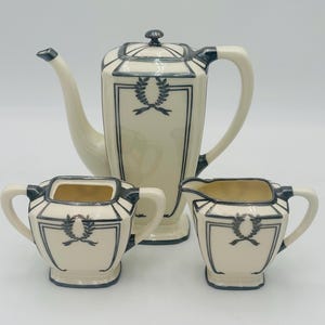 May include: A vintage cream-colored tea set with black and silver accents. The teapot, creamer, and sugar bowl feature a geometric design with a laurel wreath motif.