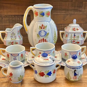 May include: A white ceramic tea set with floral and geometric designs. The set includes a pitcher, creamer, sugar bowl, cups, saucers, and small lidded pots. The designs feature red, blue, and green floral patterns.