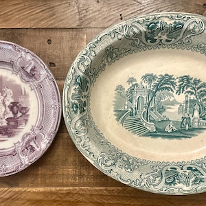 May include: Two antique ceramic plates with intricate floral designs. The larger plate is oval shaped and features a green and white landscape scene. The smaller plate is round and features a purple and white landscape scene.