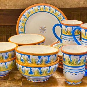 May include: A collection of ceramic tableware with a vibrant design. The set includes bowls, plates, and mugs, all featuring a white base with blue, yellow, and orange decorative patterns. The mugs have blue handles.