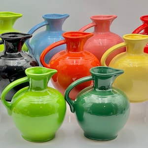 May include: A collection of colorful ceramic pitchers in various shades, including green, yellow, red, orange, pink, blue, and black. Each pitcher has a rounded body, a handle, and a spout. The pitchers are arranged on clear stands.