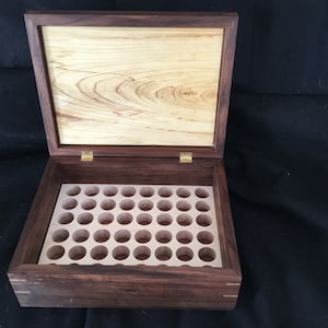 以下が含まれることがあります： ヒンジ付きの蓋が付いた開いた木製の箱。箱は濃い茶色の木材でできており、内側と蓋は明るい色の木材です。内部には複数の円形の切り抜きがあります。蓋が開いており、内部が見えます。
