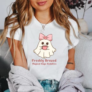 May include: White t-shirt with a cartoon ghost wearing a pink bow and holding a pink cup. The text "Freshly Brewed Magical Rage Sedative" is printed below the ghost.