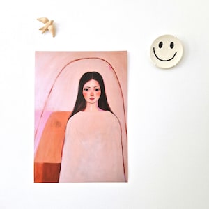 Op de afbeelding: Een kunstdruk met een vrouw met lang zwart haar en een wit kledingstuk. Het schilderij heeft een zacht, roze en perzikkleurig kleurenpalet. Een kleine houten vogel en een smiley-bord zijn ook zichtbaar.