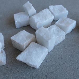 May include: A collection of white, roughly-cut quartz stones. The stones are rectangular and have a slightly rough texture. The stones are scattered on a gray surface. The quartz stones are a bright white color.